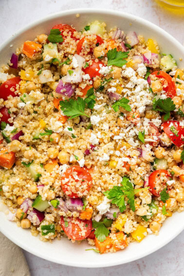 A mixed quinoa chickpea salad with tomatoes, cucumbers, feta, and parsley, in a white bowl.