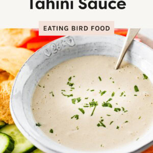Bowl of tahini sauce topped with parsley, surrounded by fresh veggies and pita chips.