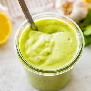 Glass jar filled with avocado dressing, garnished with parsley, lemon, and garlic in the background.