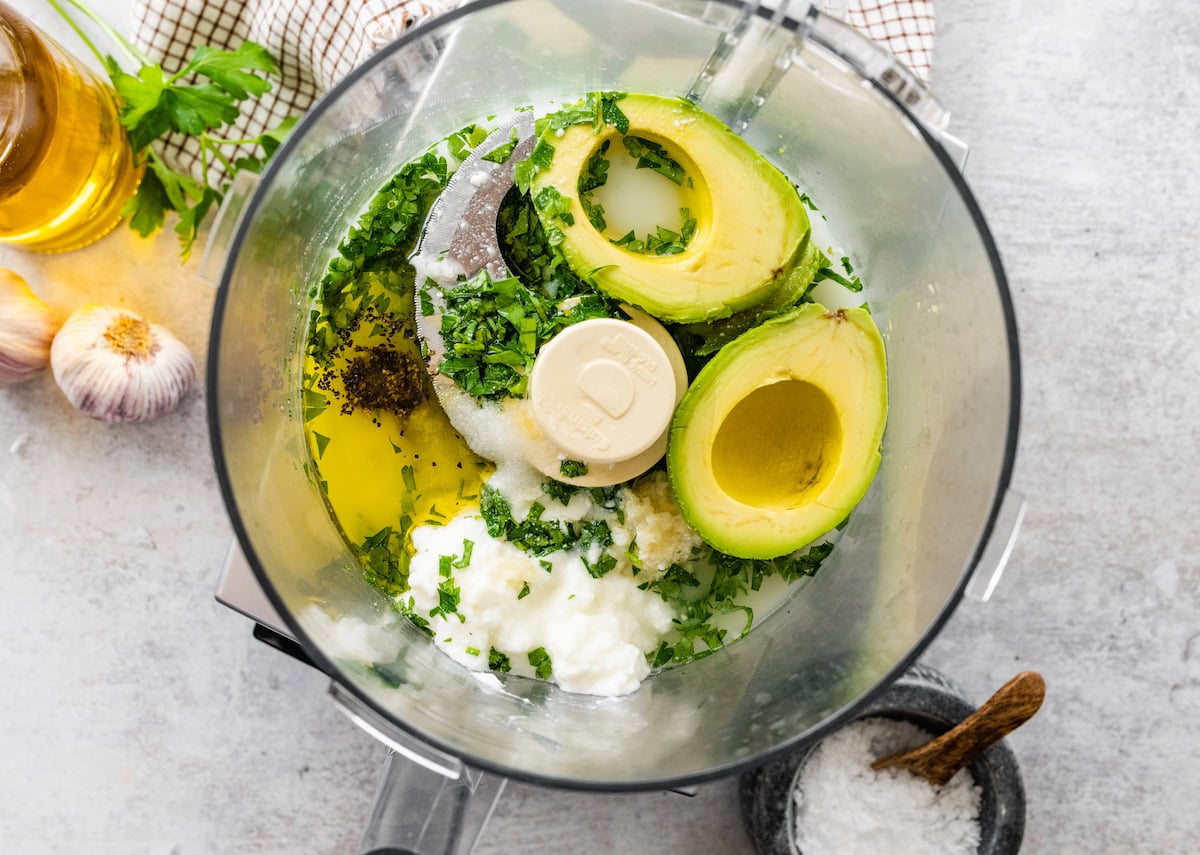 Ingredients for avocado dressing in a food processor, including avocado, Greek yogurt, lemon, and parsley.