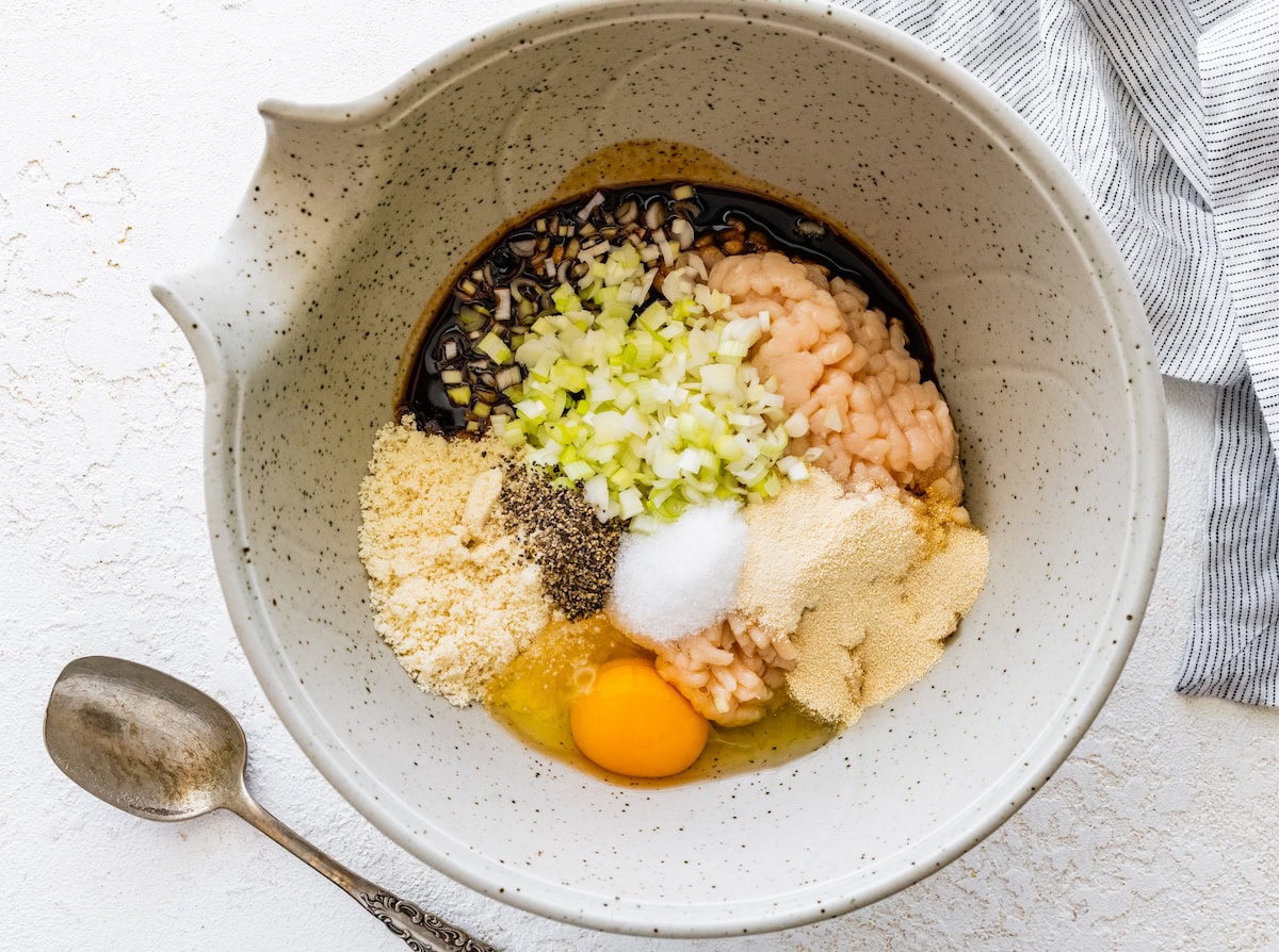 Unmixed meatball ingredients in a bowl including ground chicken, egg, almond flour, onion, and sauce.