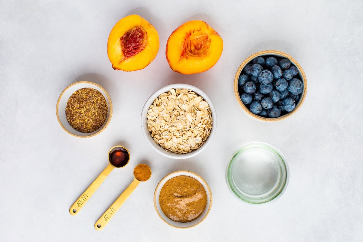 Blueberry Peach Oatmeal - Loaded with fiber, healthy fat and protein this delicious bowl of oats will keep you feeling full and energized all morning. Ingredients measured out to make blueberry peach oatmeal: blueberries, water, peaches, oats, almond butter, cinnamon, vanilla and flaxseed.