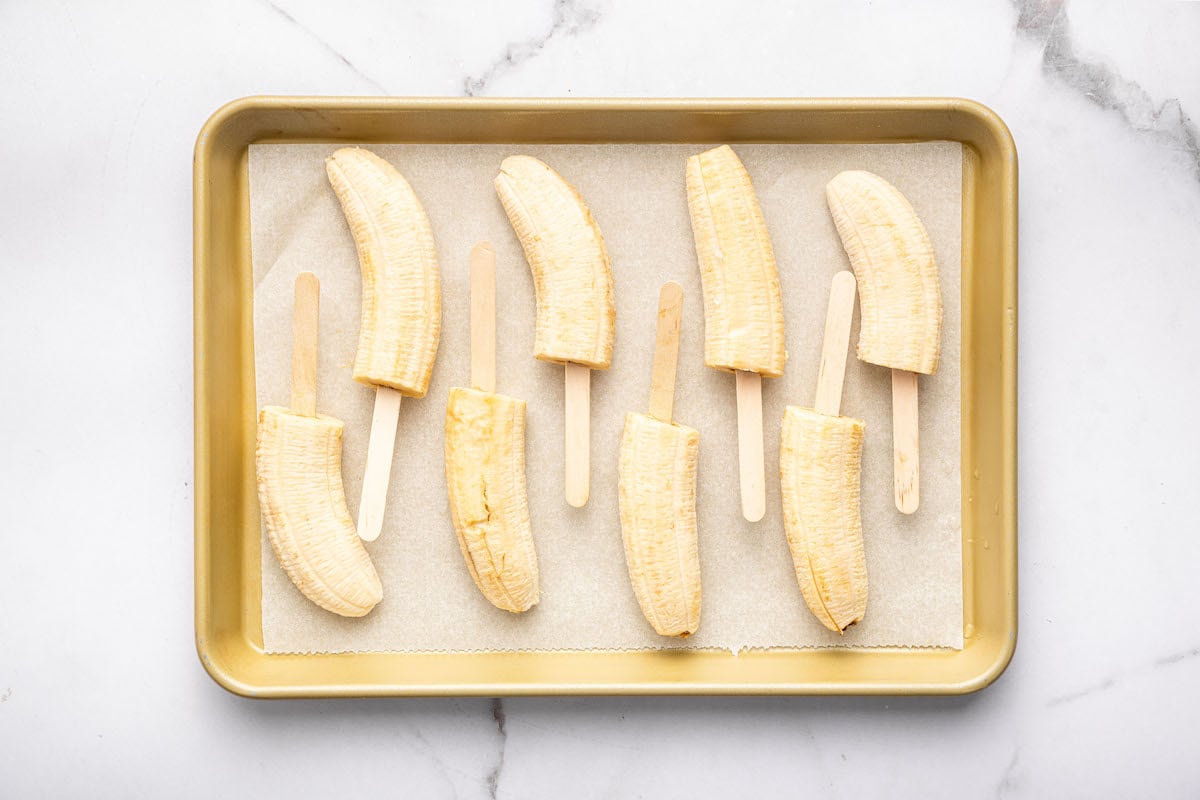 Eight banana halves on wooden sticks arranged on a parchment-lined baking tray, prepared for freezing or dipping, set on a white marble surface.