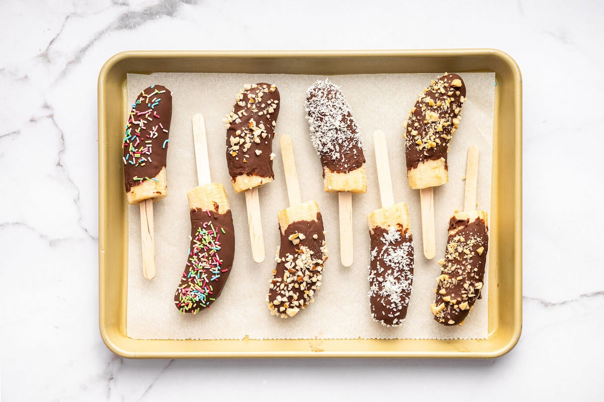 Tray of chocolate-dipped banana halves on sticks, topped with rainbow sprinkles, chopped nuts, and shredded coconut, arranged on parchment paper.