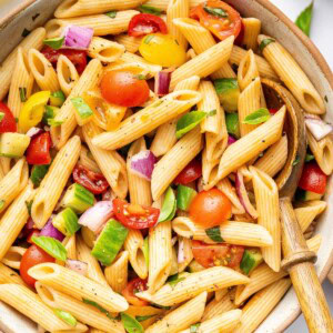 A pasta salad in serving bowl with cherry tomatoes, cucumber, red onion and fresh basil. There is a wooden serving spoon in the bowl.