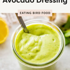 Glass jar filled with thick, creamy avocado dressing with a spoon inside, surrounded by fresh ingredients like lemon and garlic.