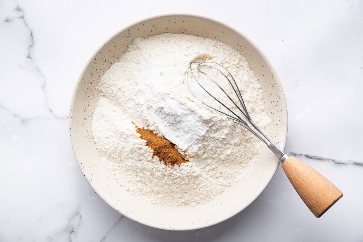 Dry ingredients with flour, baking powder, and cinnamon in a mixing bowl with a whisk.