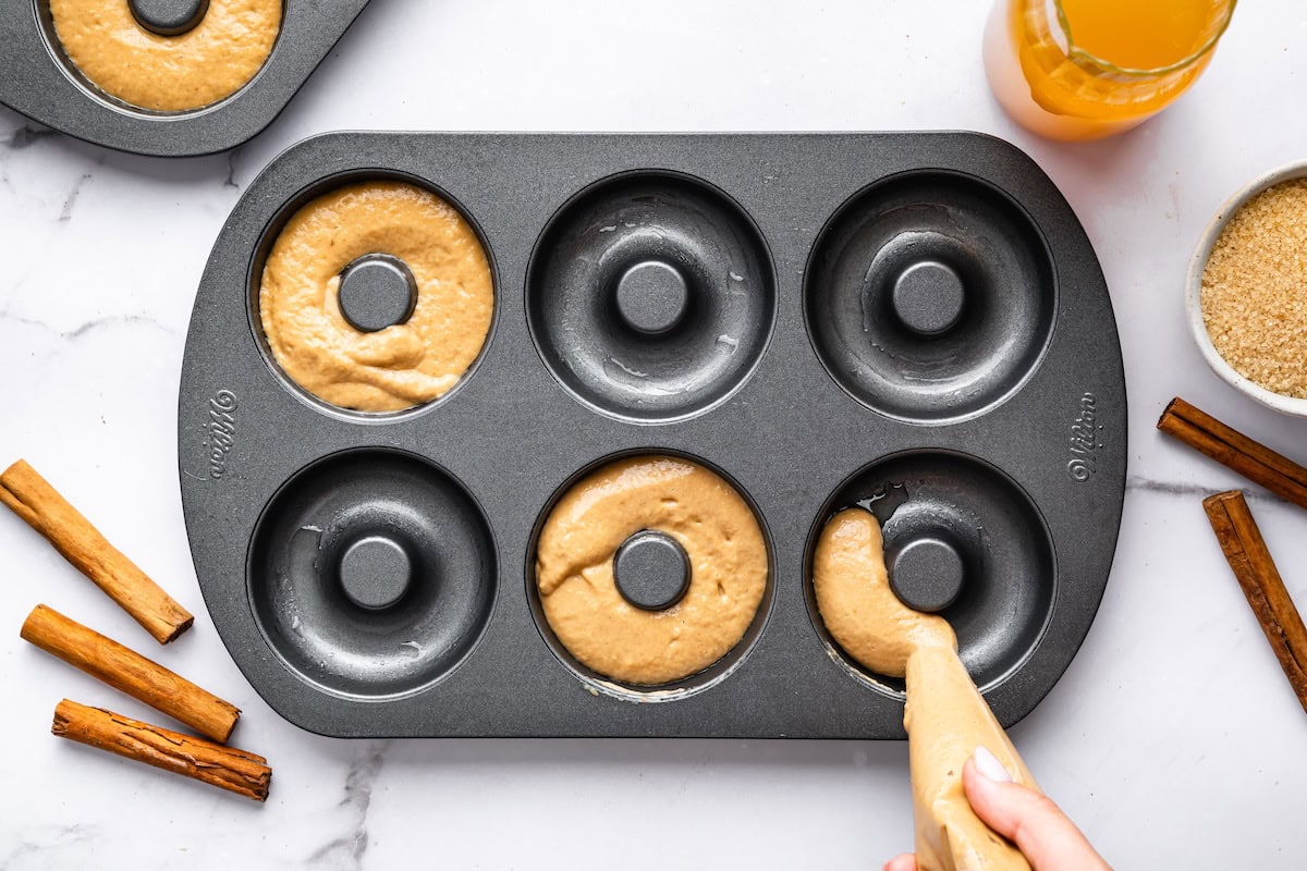 Donut batter being piped into a donut pan cavity, surrounded by cinnamon sticks and a glass.