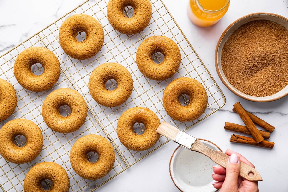 Plain baked donuts cooling on a wire rack while being brushed with melted butter before coating in cinnamon sugar.