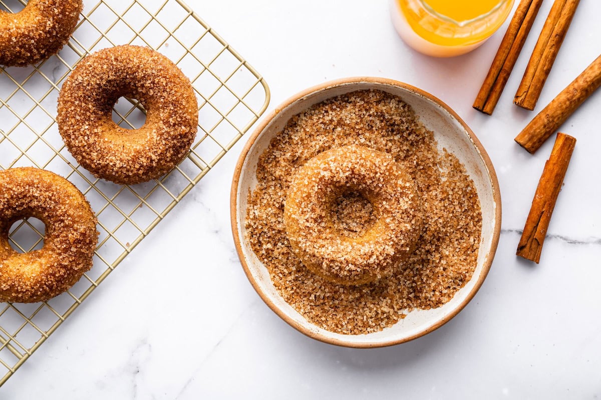 A donut being coated in a bowl of cinnamon sugar with more donuts cooling on a wire rack.