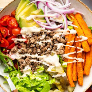 Assembled burger bowl with lettuce, tomato, onion, pickles, avocado, fries, and beef all drizzled with Big Mac sauce.