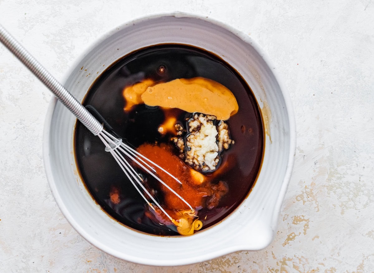 White mixing bowl with peanut sauce ingredients including soy sauce, peanut butter, and garlic.