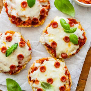 English muffin pizzas topped with melted cheese, mini pepperoni and basil leaves, served on parchment.