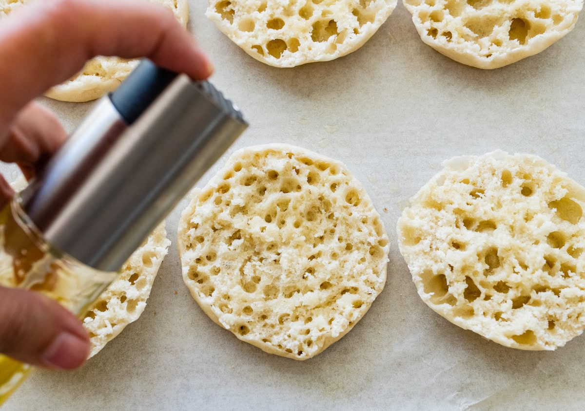 Spraying English muffin halves with olive oil before adding toppings.