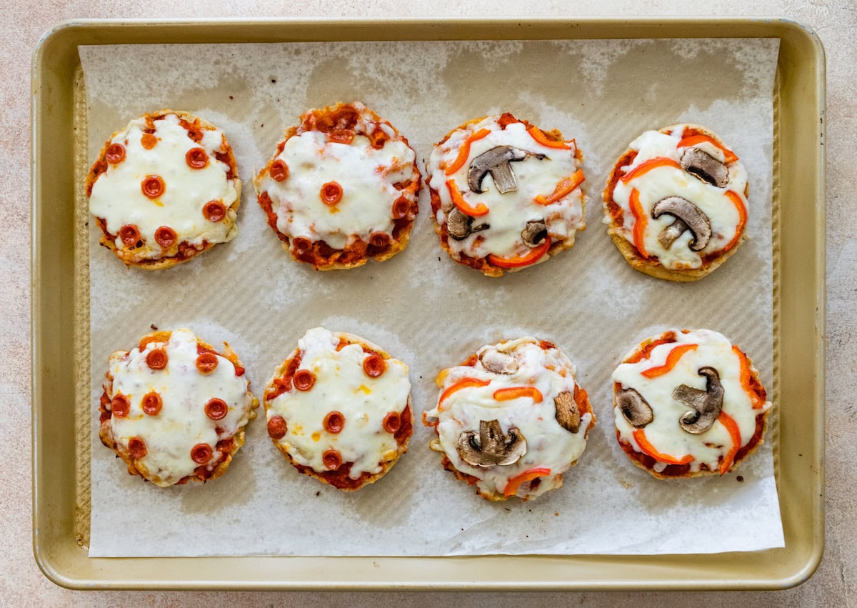 Baked English muffin pizzas on a baking sheet with melted mozzarella and toppings.