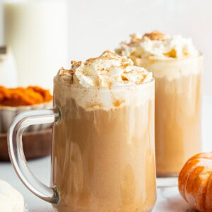 Two glass mugs filled with pumpkin spice lattes topped with whipped cream and cinnamon, set among pumpkins, milk, and fall spices.