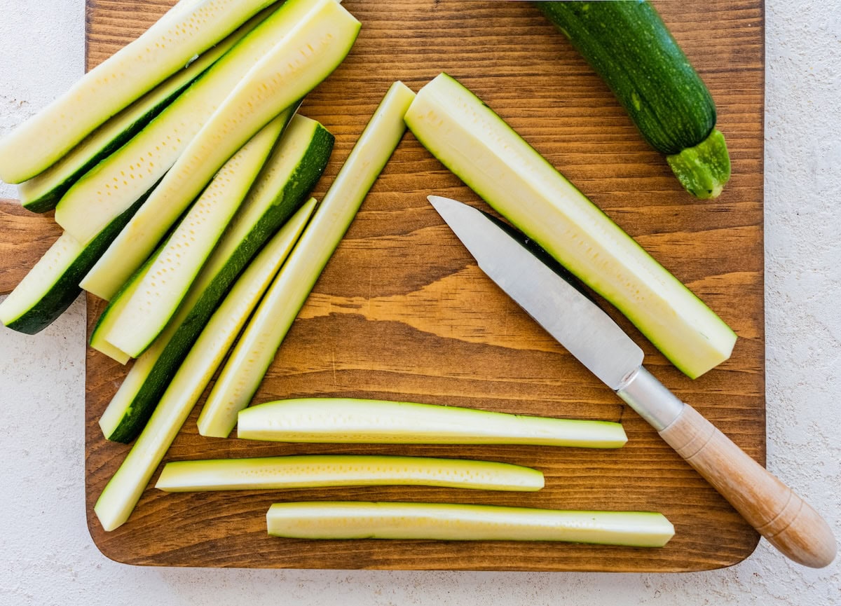 Fresh zucchini sliced into sticks on a wooden cutting board with a knife.