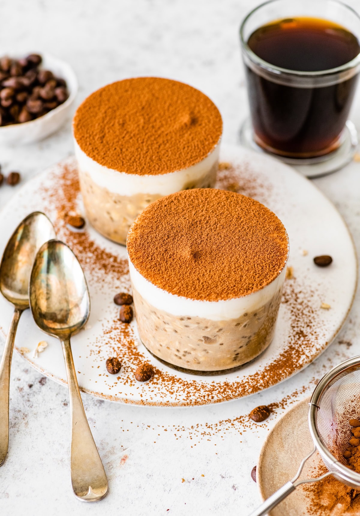 Two jars of tiramisu overnight oats topped with cocoa powder, served with coffee and vintage spoons.