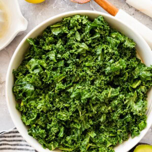 Massaged kale salad in a large white serving bowl with a lemon and wooden salad servers beside it.