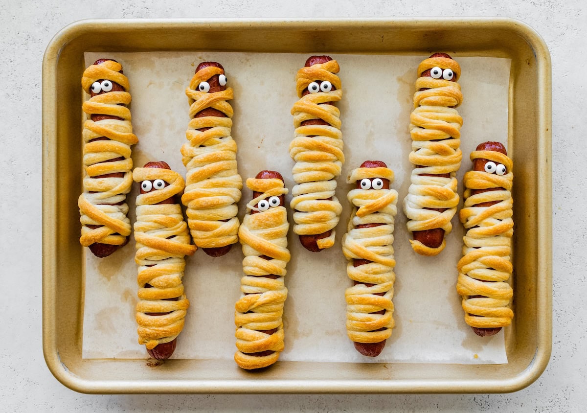 Freshly baked mummy hot dogs with candy eyes on a sheet pan, wrapped in crescent roll dough for Halloween.