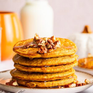 Five pumpkin protein pancakes stacked on one another, served on a plate with chopped pecans and maple syrup.