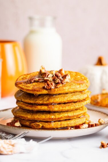 Five pumpkin protein pancakes stacked on one another, served on a plate with chopped pecans and maple syrup.