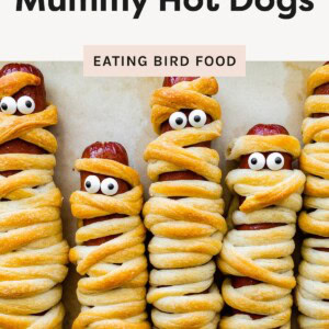 Five mummy hot dogs wrapped in golden crescent roll dough with candy eyes, lined up side by side on parchment paper