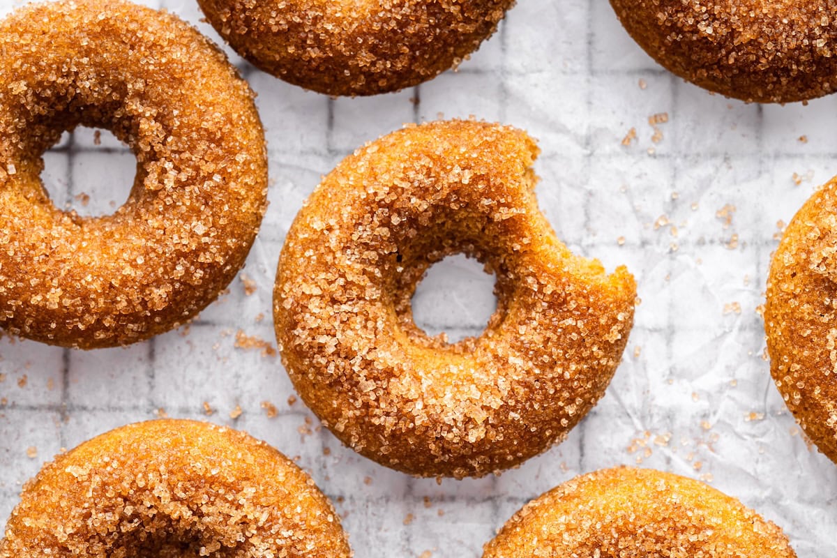 A sugar coated baked donut on parchment paper on a cooling rack with a bite taken out of it near more donuts.