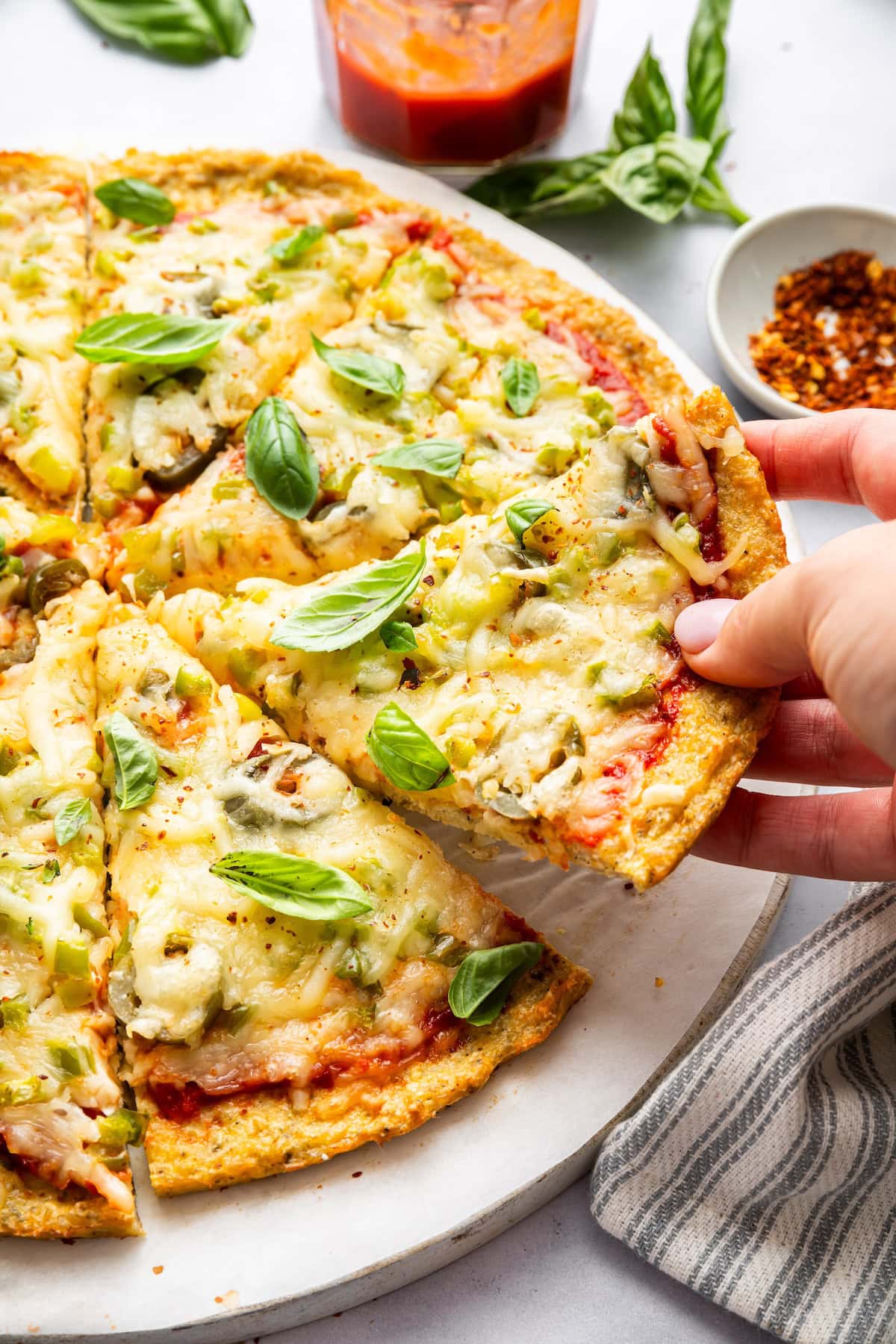 A hand grabbing a slice of a chicken pizza crust topped with fresh basil.