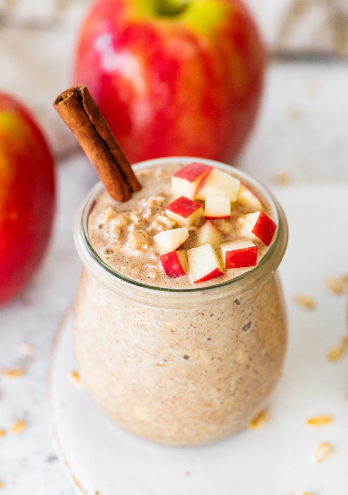 Two jars of apple cinnamon overnight oats on a white plate topped with chopped apples and cinnamon sticks with oats scattered around.