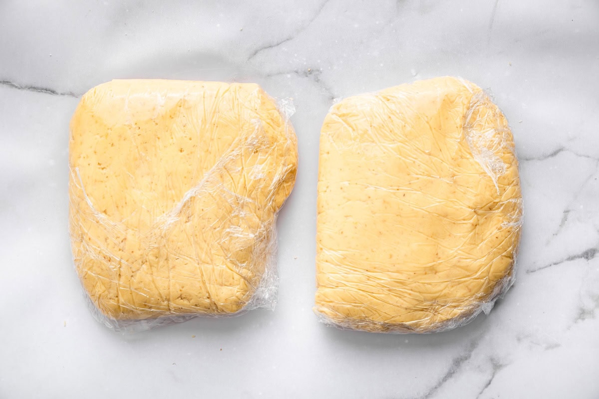 Two flattened discs of sugar cookie dough wrapped tightly in plastic wrap resting on a white marble surface. The dough is smooth, pale yellow, and ready to be chilled before rolling.