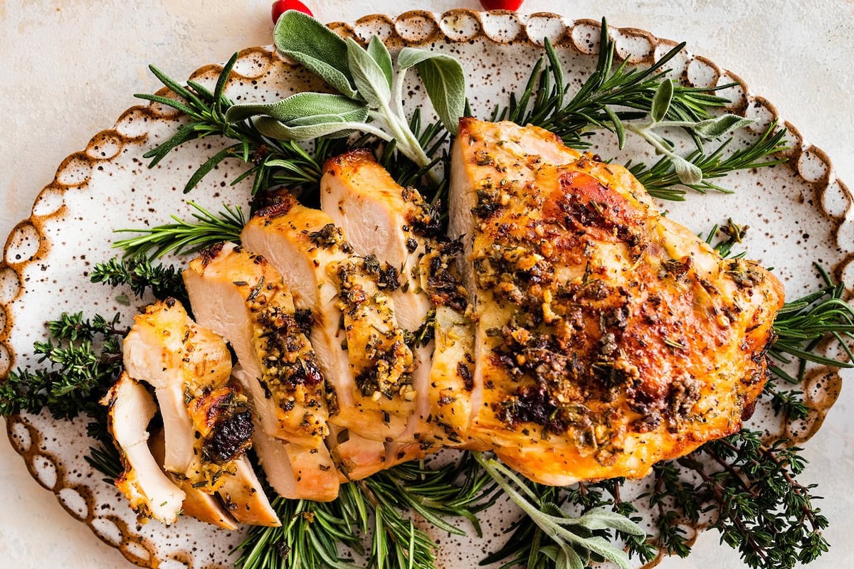 An air fryer turkey breast sliced sitting on a bed of fresh herbs and cranberries on a white platter.