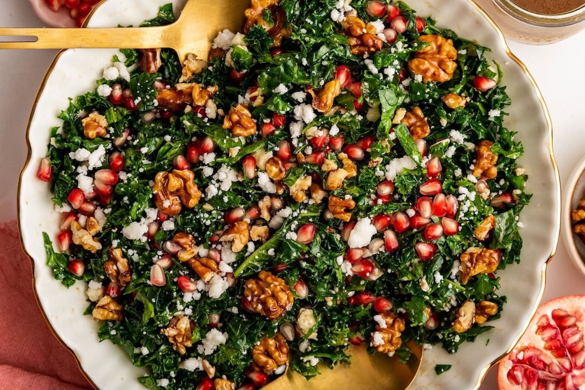 A kale salad topped with fresh pomegranate, goat cheese and candied walnuts. The salad is served in a white bowl with gold salad spoons.