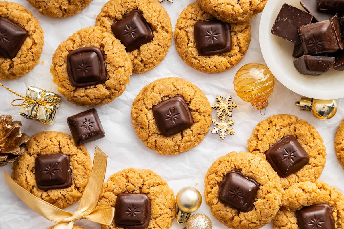 Peanut butter blossom cookies arranged on white parchment with small gold ornaments, and extra chocolate pieces scattered around.