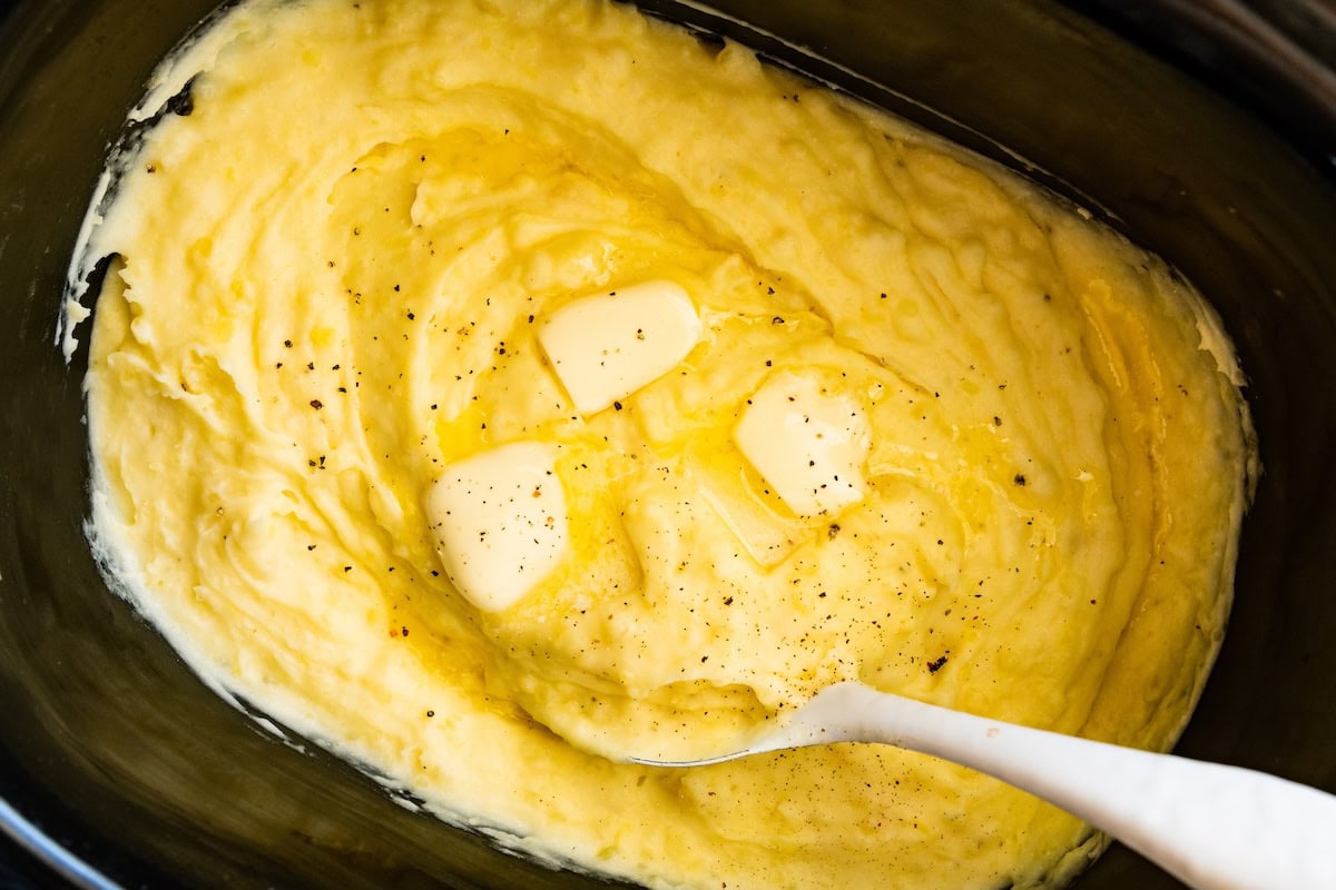 Mashed potatoes in a slow cooker with slices of butter, seasonings, and a spatula.