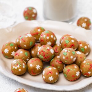 A cream-colored plate piled with sugar cookie protein balls decorated with festive sprinkles, with a glass of milk in the background.