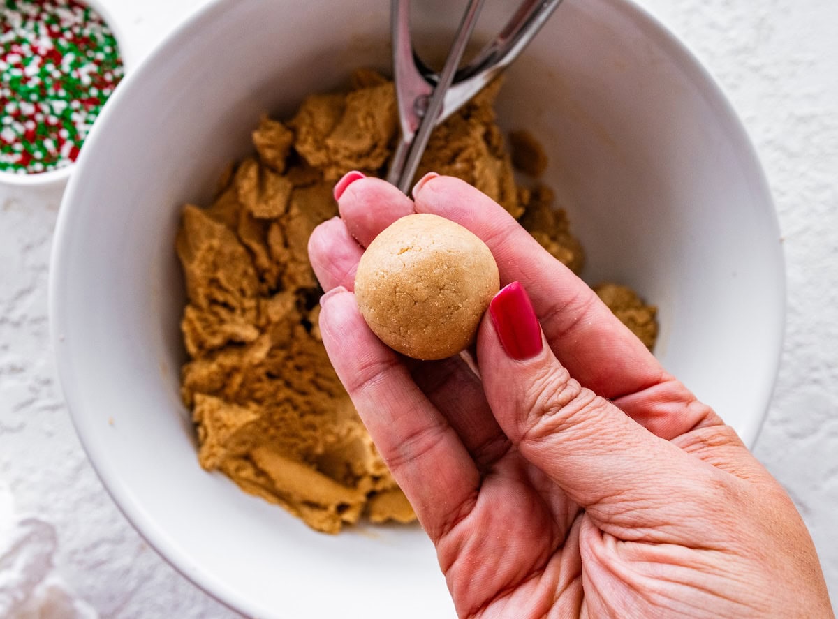 form balls A hand holding a small, round piece of sugar cookie protein ball dough over a bowl of dough, just before rolling in sprinkles.