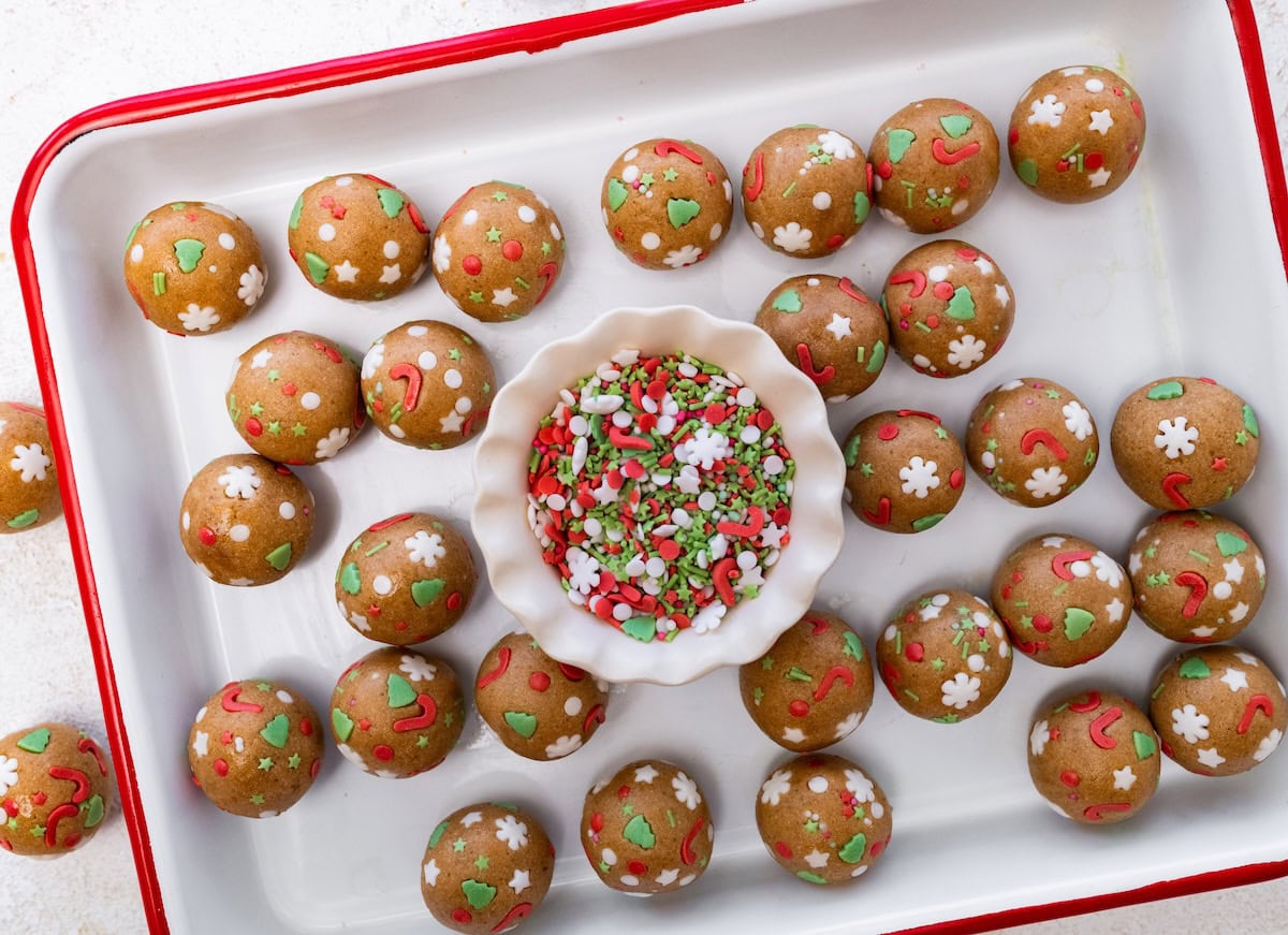 roll balls in sprinkles A rectangular tray lined with sugar cookie protein balls surrounding a small dish of red, white, and green sprinkles in the center.