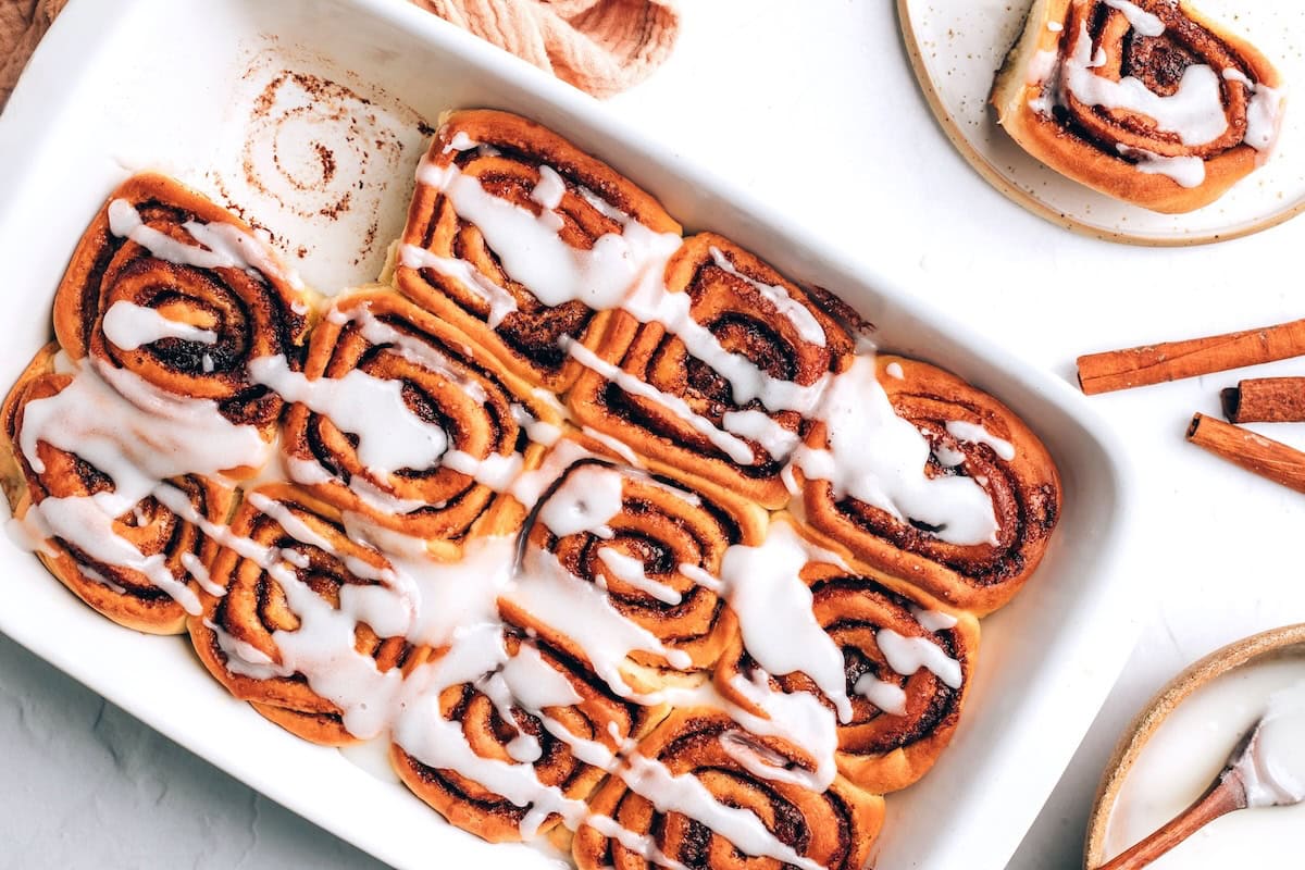 An overhead photo of vegan cinnamon rolls in a baking dish. A roll has been removed from a corner.
