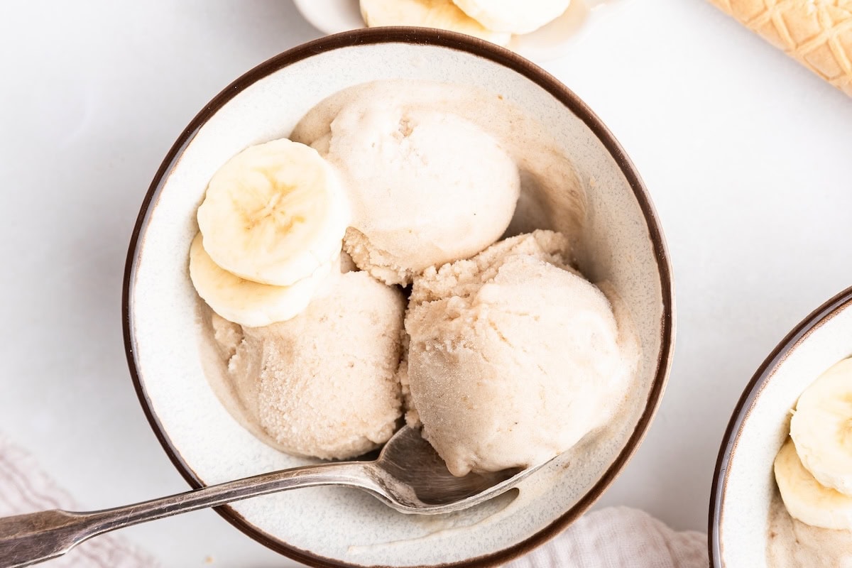 Three scoops of banana ice cream in a bowl with a metal spoon and two slices of banana.