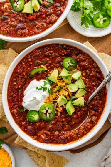 A bowl of slow cooked bean and beef chili topped with avocado, cheese, sour cream, and jalapeno slices.