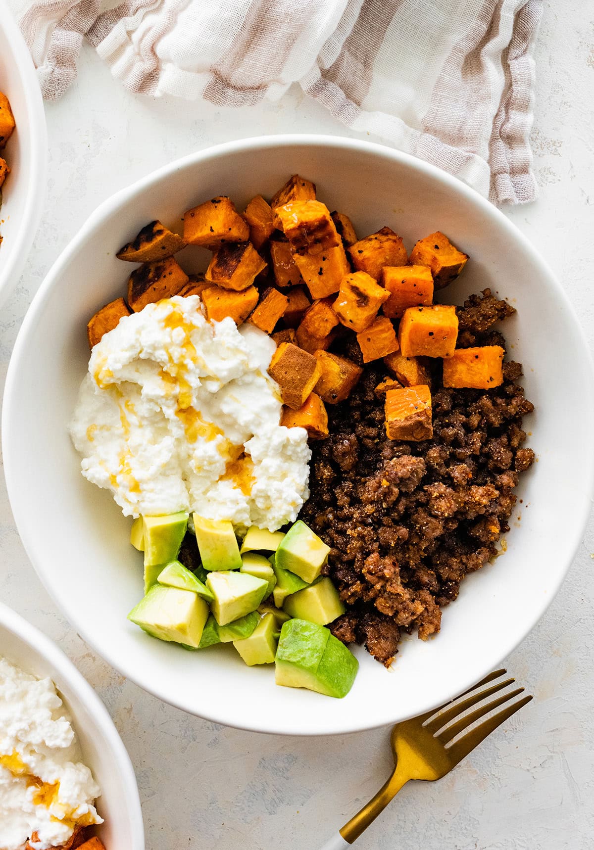 Ground beef in a white bowl with roasted sweet potatoes, cottage cheese, avocado chunks and a drizzle of hot honey.