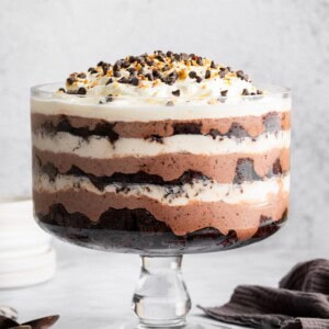 Chocolate Trifle with layers of chocolate pudding, brownie cubs and whipped topping in a glass trifle dish. The trifle is topped with chocolate chips and chopped nuts.
