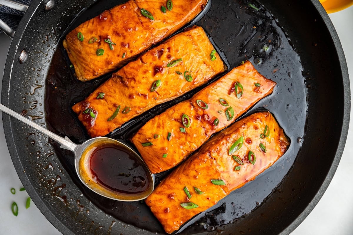 Four honey glazed salmon filets garnished with fresh chives in a skillet with a metal spoon that contains some of the honey based sauce in it.