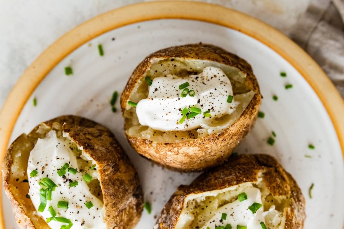 Three baked potatoes split open on a plate with sour cream, fresh chives, and seasoning.