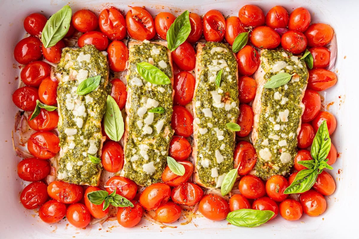 Four salmon filets covered in pesto and surrounded by cherry tomatoes in a baking dish.