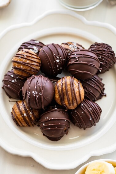 Banana bread protein balls on a plate. Half are dipped in chocolate and half are drizzled with chocolate.