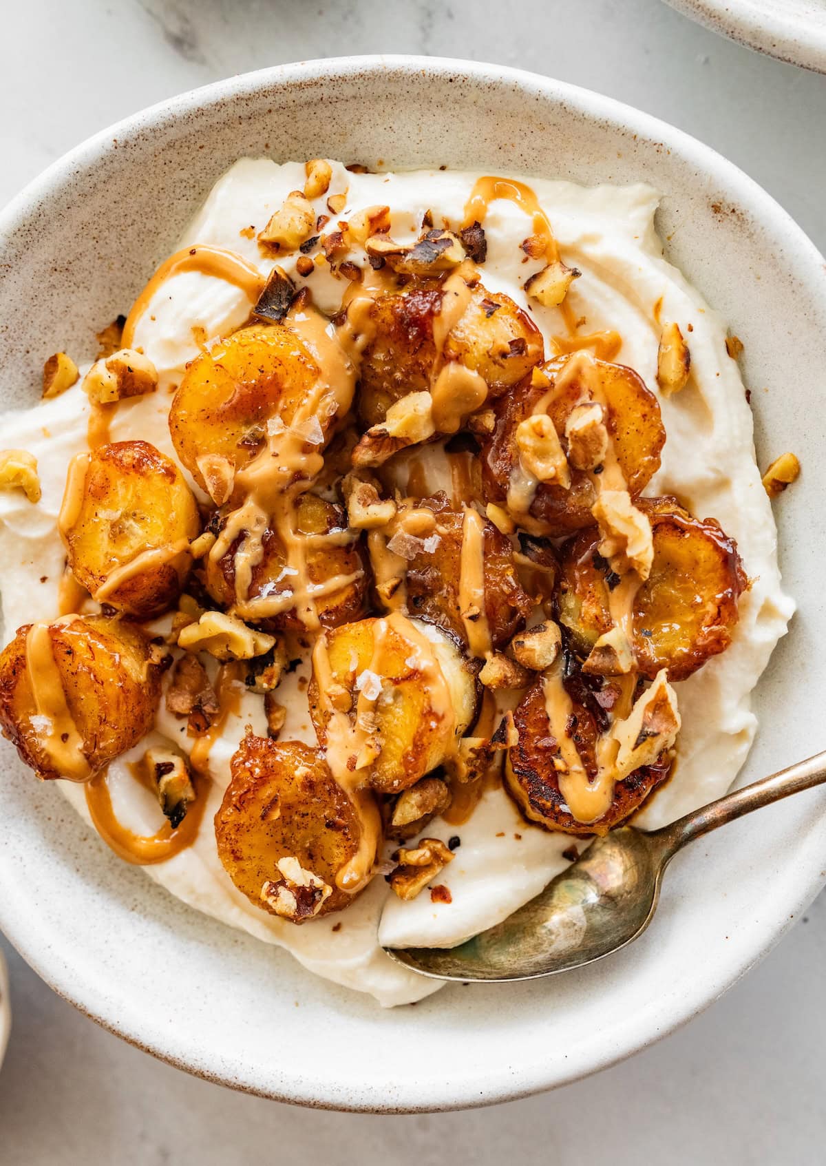 A caramelized banana yogurt bowl in a speckled bowl topped with a drizzle of peanut butter and chopped walnuts.