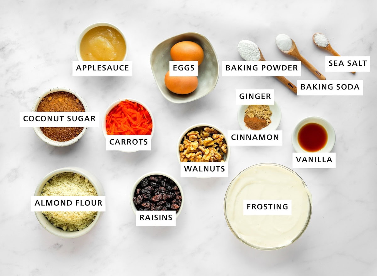 Ingredients measured out to make carrot cake bars: almond flour, coconut sugar, applesauce, carrots, raisins, eggs, walnuts, ginger, cinnamon, homemade frosting, baking soda, baking powder, vanilla and sea salt.