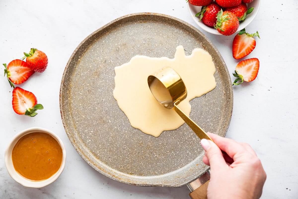 A womans hand uses a measuring cup to pour protein crepe batter on a skillet.